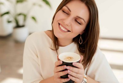 High angle of cheerful young female smiling with closed eyes and enjoying smell of freshly brewed coffee while spending time in cafe in morning